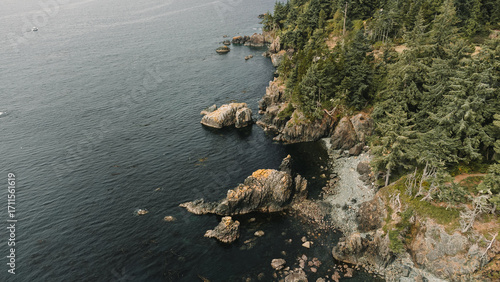 An aerial, drone shot of a white lighthouse with a red top, situated on a rocky cliff on a coastline. The dark ocean water surrounds the rugged shoreline, which is lined with evergreen trees