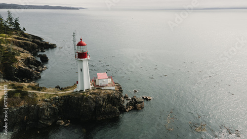 An aerial, drone shot of a white lighthouse with a red top, situated on a rocky cliff on a coastline. The dark ocean water surrounds the rugged shoreline, which is lined with evergreen trees