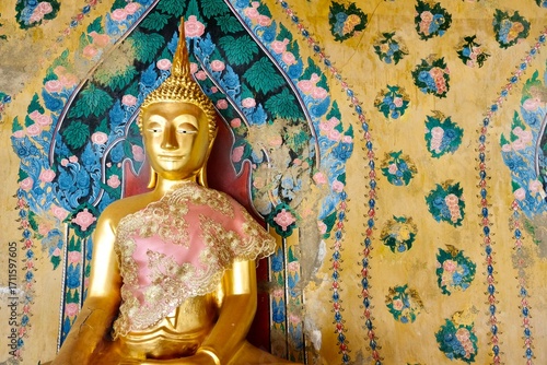 Gold Buddha statue clothed in traditional Thai silk garments with ancient painted backdrop at Wat Arun, the Temple of Dawn in Bangkok, Thailand
