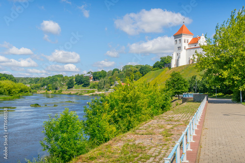 Riverbank pathway along the Neman River in Hrodna, Belarus, with the historic Old Grodno Castle on a grassy hillside, during a bright summer day.
