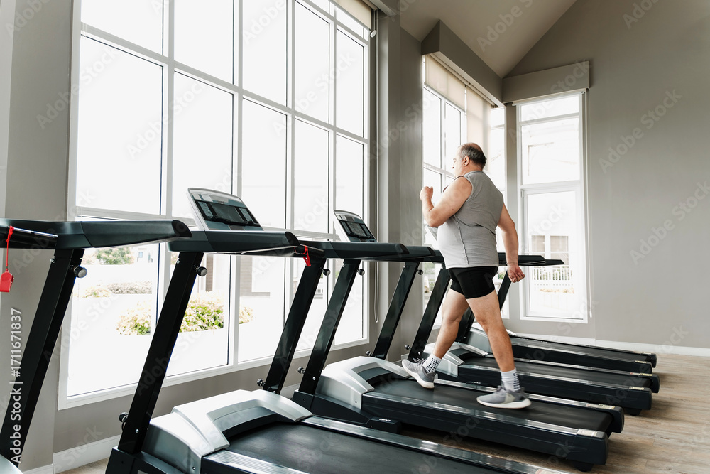 Fototapeta premium Man walks on treadmill in bright, spacious gym with large windows. Scene shows cardio fitness, wellness, wellbeing, motivation, performance, coaching, strength, running, jogging, and rehabilitation.