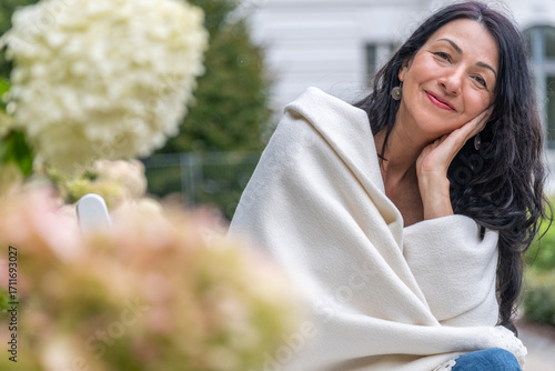 Elderly woman wrapped in a soft blanket, sitting in a garden surrounded by blooming flowers, radiating elegance and wisdom while enjoying a moment of natural beauty