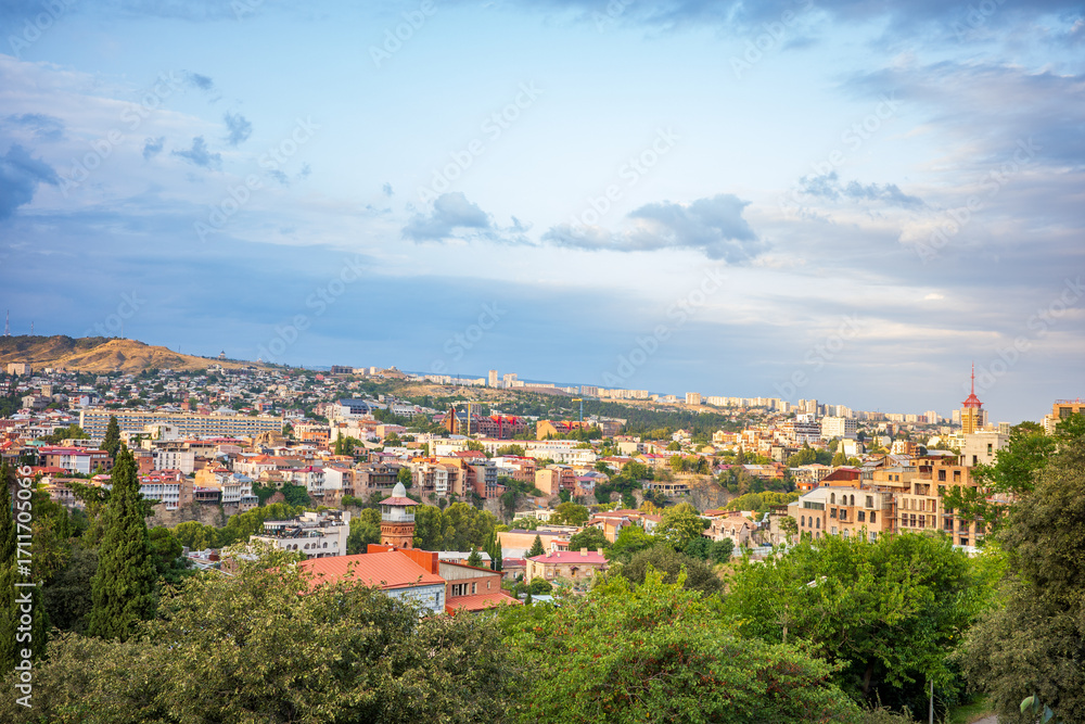 Fototapeta premium Stunning panoramic view of Tbilisi, Georgia, captured at sunset. The city’s colorful rooftops, lush greenery, and rolling hills blend beautifully under a glowing sky of Georgia’s capital town