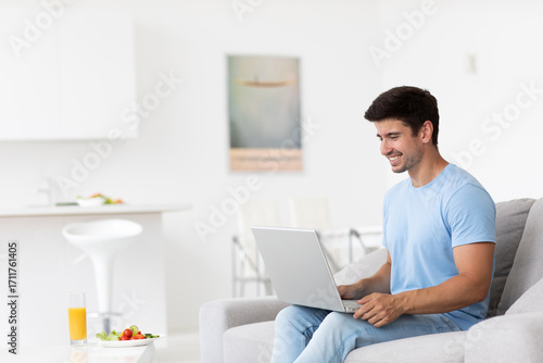 Cheerful guy is comfortably seated on a sofa in his own room with a laptop, smiling