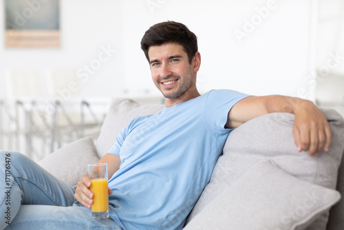 Handsome young man sitting on the sofa in the living room with juice, relaxing and eating healthy, looking