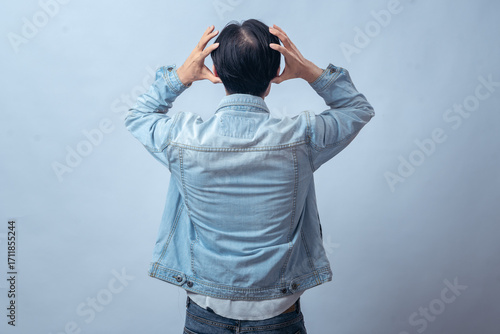 Asian man in denim jacket seen from behind, holding head with both hands in frustration, standing in studio against plain light background, expressing stress, tension, and emotional struggle