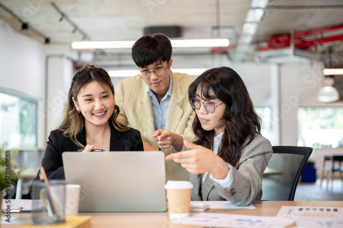 Asian businesswoman talking with glasses coworkers while looking at laptop on wooden table in office