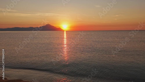 Breathtaking aerial drone capture of a tranquil sunset over a peaceful sea, with the sun dipping behind a distant mountain range. The sky glows in rich shades of orange and yellow, transitioning into 