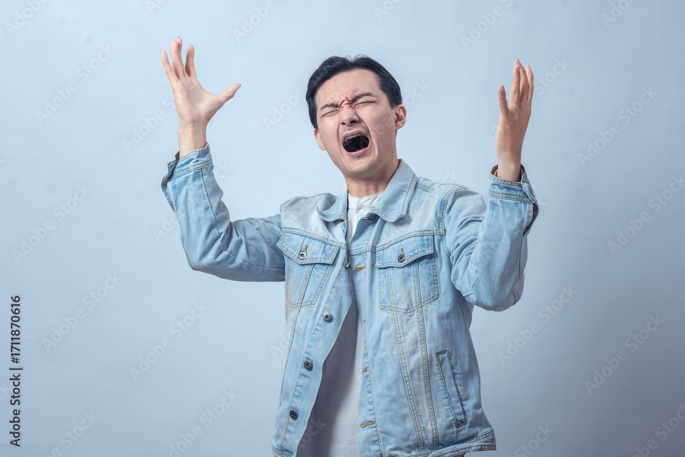 Naklejka premium Asian man in denim jacket screaming with both hands raised, expressing anger, frustration, sadness, or despair on a plain studio background, dramatic emotional reaction
