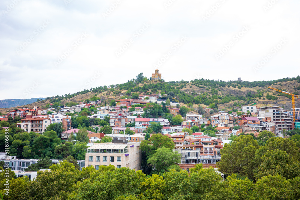 Fototapeta premium Colorful hillside neighborhood in Tbilisi, Georgia, with traditional houses, modern buildings, and a historic church on the hilltop. This scenic view captures the blend of old and new architecture