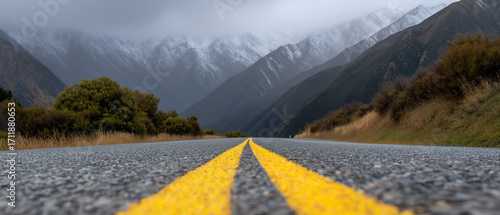 Mountain road with yellow dividing lines stretches into foggy, snow-capped peaks under overcast skies, flanked by bushes and grassy hillsides