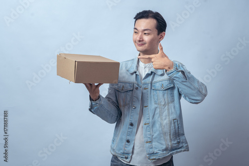 Asian man in denim jacket holding cardboard box on one hand and pointing at it with smile against plain studio background, symbolizing delivery, shipping, product promotion, or ecommerce service