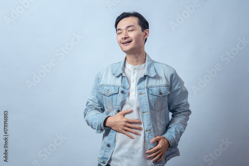 Asian man in denim jacket touching stomach with pained face, standing in studio against plain light background, expressing stomachache, discomfort, and sick feeling with casual fashion style