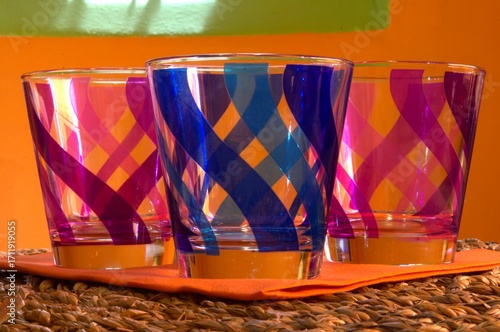 Three colored glasses placed on a woven straw shelf and with an orange wall in the background