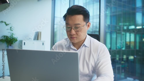 Serious Asian male in white shirt and glasses sitting at desk in bright office using laptop. Reading document or reviewing data. Concentrating on numbers or checking client presentation.