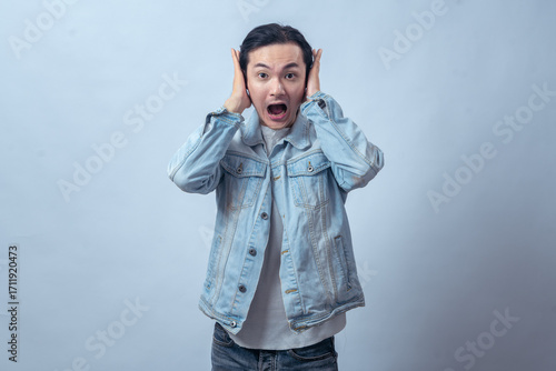 Asian man in denim jacket covering ears with hands and shouting in shock, standing in studio against plain light background, showing surprised expression and emotional reaction