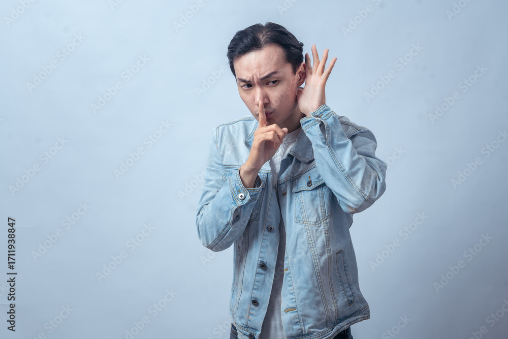 Fototapeta premium Asian man in denim jacket making silence gesture with finger on lips and hand cupped to ear, listening carefully, serious expression, concept of secret, hush, or confidential talk