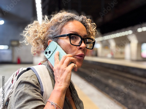 Fotografi Middle-aged woman on phone in underground train station, worried and waiting for