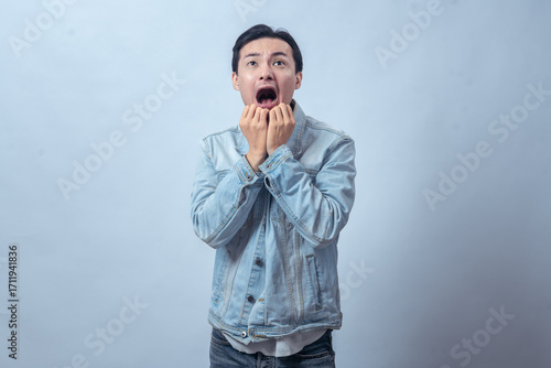 Asian man in denim jacket biting nails with anxious face and tense body language, standing in studio against plain light background, expressing nervousness, stress, and fear