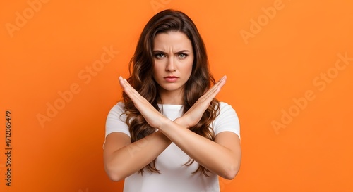 Serious young woman with a determined expression making a stop gesture with her crossed arms, showing rejection and denial on an orange background