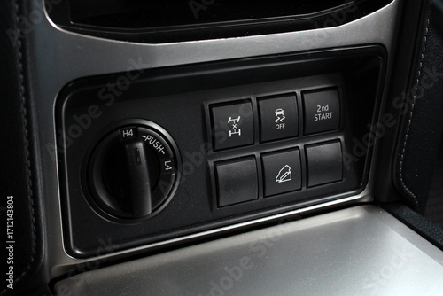 Close-up of black SUV control panel with rotary dial and rectangular buttons for off-road driving, traction, and stability functions, integrated into silver-trimmed dashboard design.