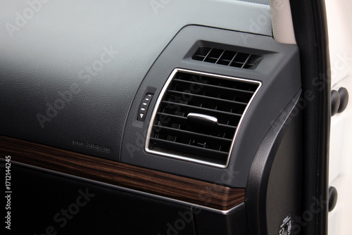 Close-up of car dashboard with black textured surface, wood trim detail, chrome-framed air vent, adjustment slider, and integrated SRS airbag marking on passenger side.