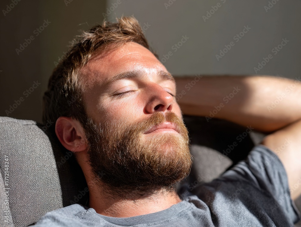 Obraz premium A serene man with a beard is seen relaxing or sleeping on a sofa, bathed in warm sunlight.