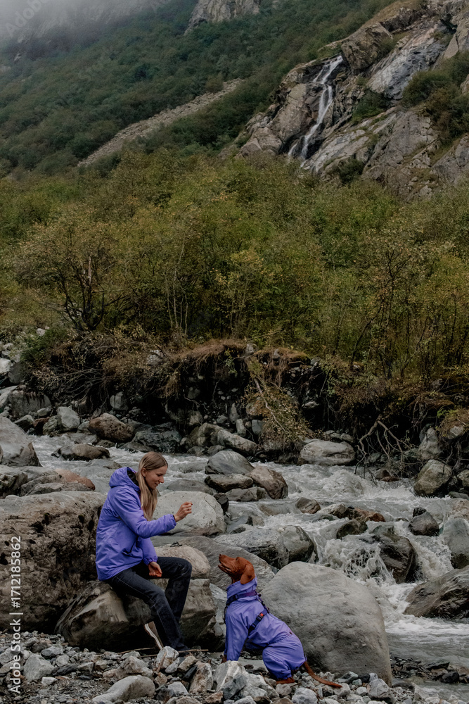 Fototapeta premium Young Caucasian man and a child sit on rocks by a river in a mountainous area. Both wear purple jackets. Lush greenery surrounds the scene.
