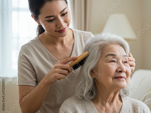 A younger Asian woman gently brushes the gray hair of an older Asian woman, who smiles serenely, conveying care and comfort in a home setting.