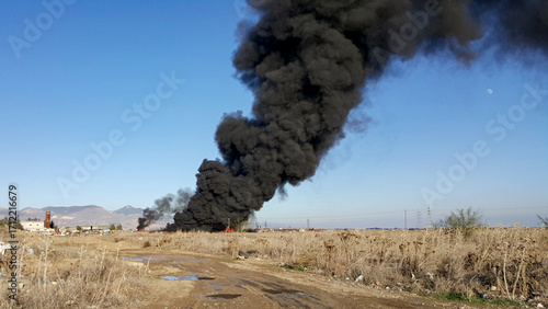 A large plume of black smoke rises from a fire in a junkyard or waste site. The image highlights the extreme environmental pollution and hazard of waste fires.