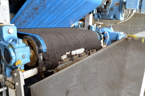 A dirty industrial belt filter press in operation, with sludge being dewatered on a moving conveyor belt, representing wastewater treatment and environmental technology.