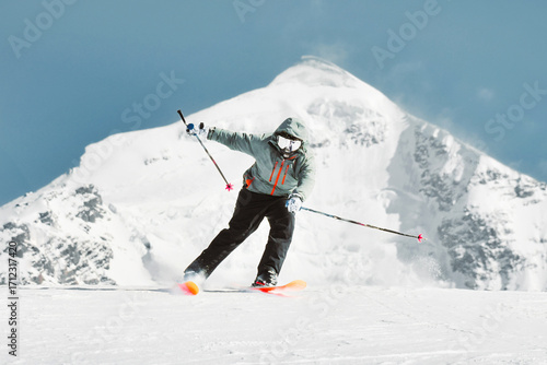 Front view skier on hilltop full speed ski downhill in sunny day in winter ski resort with mountain top background