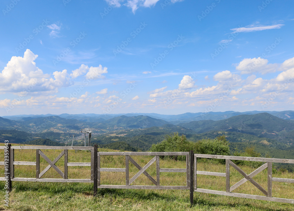 Fototapeta premium A vast mountain landscape, framed by a wooden fence, conveys the grandeur of nature. Green hills and forests, smoothly transitioning into the blue outlines of mountains on the horizon, create a sense 
