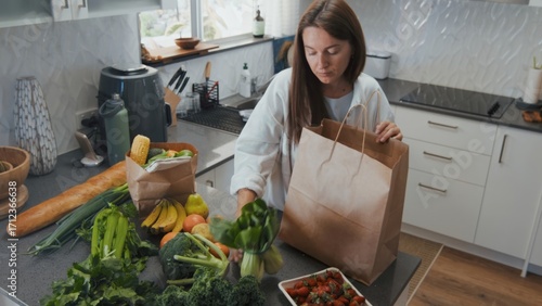 Young woman unpacks fresh groceries in cozy kitchen. Calm atmosphere highlights fruits, vegetables, and bread during daytime.