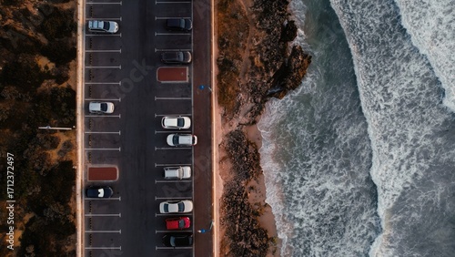 Aerial view captures multiple cars parked in vibrant colors next to serene ocean. Peaceful coastal scene evokes calmness and tranquility.