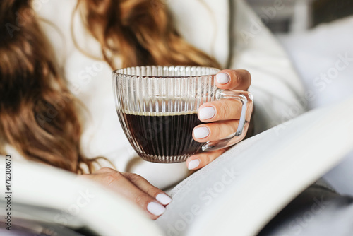 Wallpaper Mural Young woman holding glass cup of coffee and reading book. Relaxed morning routine at home. Lifestyle detail scene. Cozy home atmosphere. Hot black drink in transparent elegant cup in female hand. Torontodigital.ca