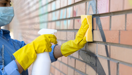 Worker in protective mask, gloves scrubs graffiti from brick wall using cleaning spray, sponge. Professional cleaning service restores building facade. Hygiene, sanitization for outdoor surfaces.