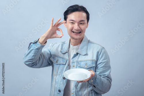 Asian man in denim jacket smiling while holding an empty white plate and giving a thumbs up gesture, isolated on plain studio background