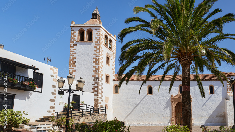 Obraz premium Historic church in Betancuria, Fuerteventura. A palm tree, whitewashed walls, and colonial architecture under a bright blue sky in this traditional Canarian village.