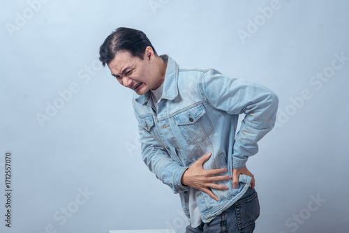 Asian man in denim jacket holding side and lower back with painful face, standing in studio against plain light background, expressing kidney pain, cramp, and body discomfort