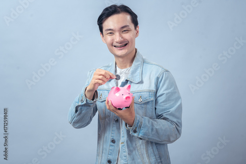 Asian man wearing a denim jacket smiling while inserting a coin into a pink piggy bank against a plain background, symbolizing saving money and financial planning