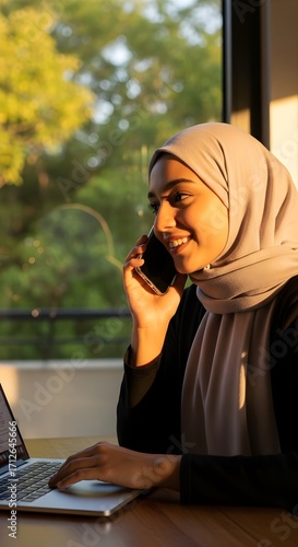 Smiling woman in hijab on phone and laptop, epitomizing modern work, communication, and multitasking in a sunlit, productive environment
