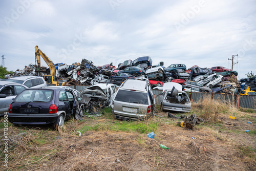 Tableau sur toile Crushed and dismantled vehicles fill a junkyard under an overcast sky, with a yellow excavator amid piles of metal