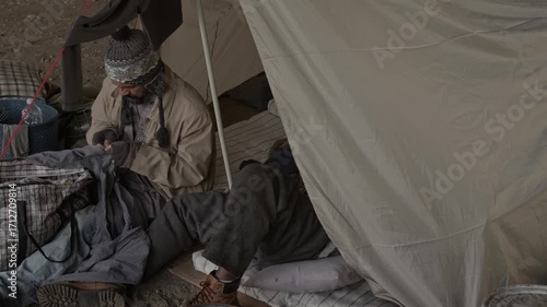 Wallpaper Mural High angle shot of concentrated bearded male vagrant mending hole in his trousers while unknown homeless man lying on mattress inside tent outdoors Torontodigital.ca