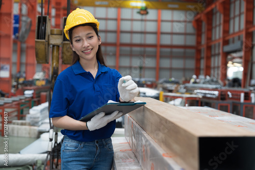 Asian woman worker check quality of square steel tubes in factory, wearing safety uniform, helmet and using clipboard. Female factory worker inspecting raw materials in storage warehouse