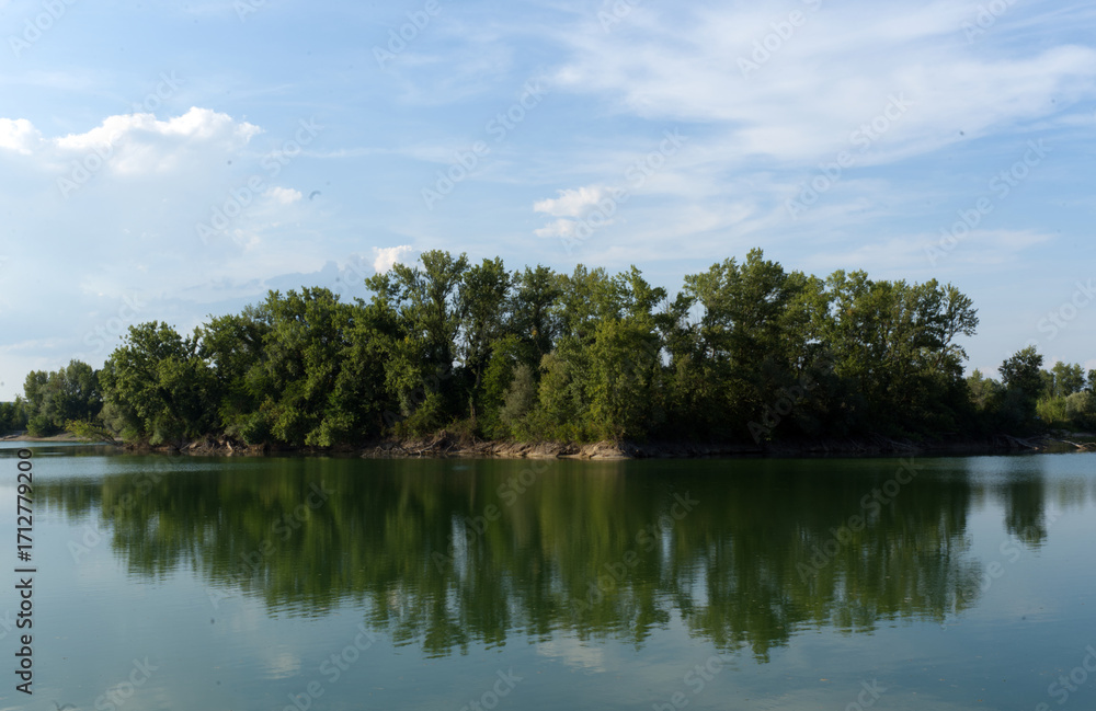 Fototapeta premium A calm body of water reflects an island densely covered with green trees under a bright blue sky with scattered clouds.