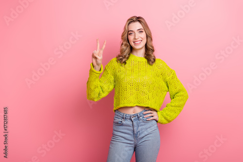 Cheerful young woman in stylish yellow knit sweater giving a peace sign gesture, smiling against a pink background