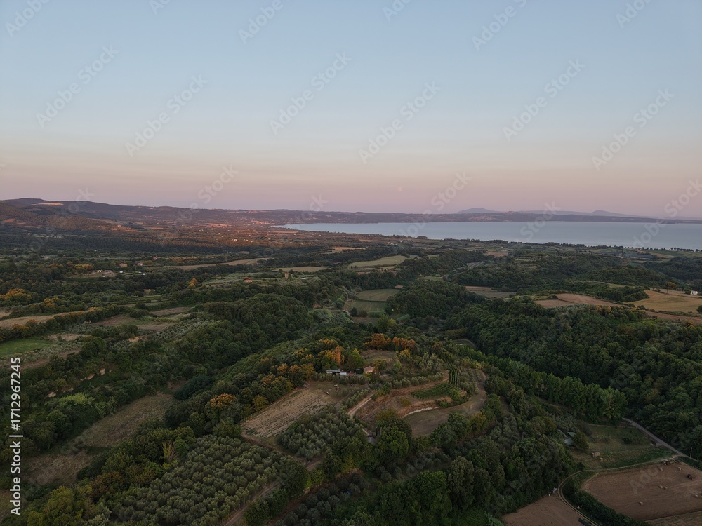 Fototapeta premium Sunset over fields and forest near Lake Bolsena, Italy Drone image showing rolling hills, farmland and forest leading to Lake Bolsena at sunset, with the full moon visible.