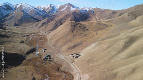 Wide aerial view of Tash Rabat caravanserai surrounded by mountain landscape, river, and snowy peaks. A historic Silk Road monument located in the Tien Shan mountains.