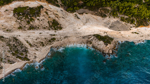 Zakynthos Greece aerial top view of rocky coastline with turquoise Ionian Sea waves crashing against limestone shore and green forest above